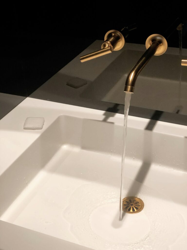 A sleek gold faucet pours water into a modern white washbasin in a dimly lit, elegant bathroom setting.